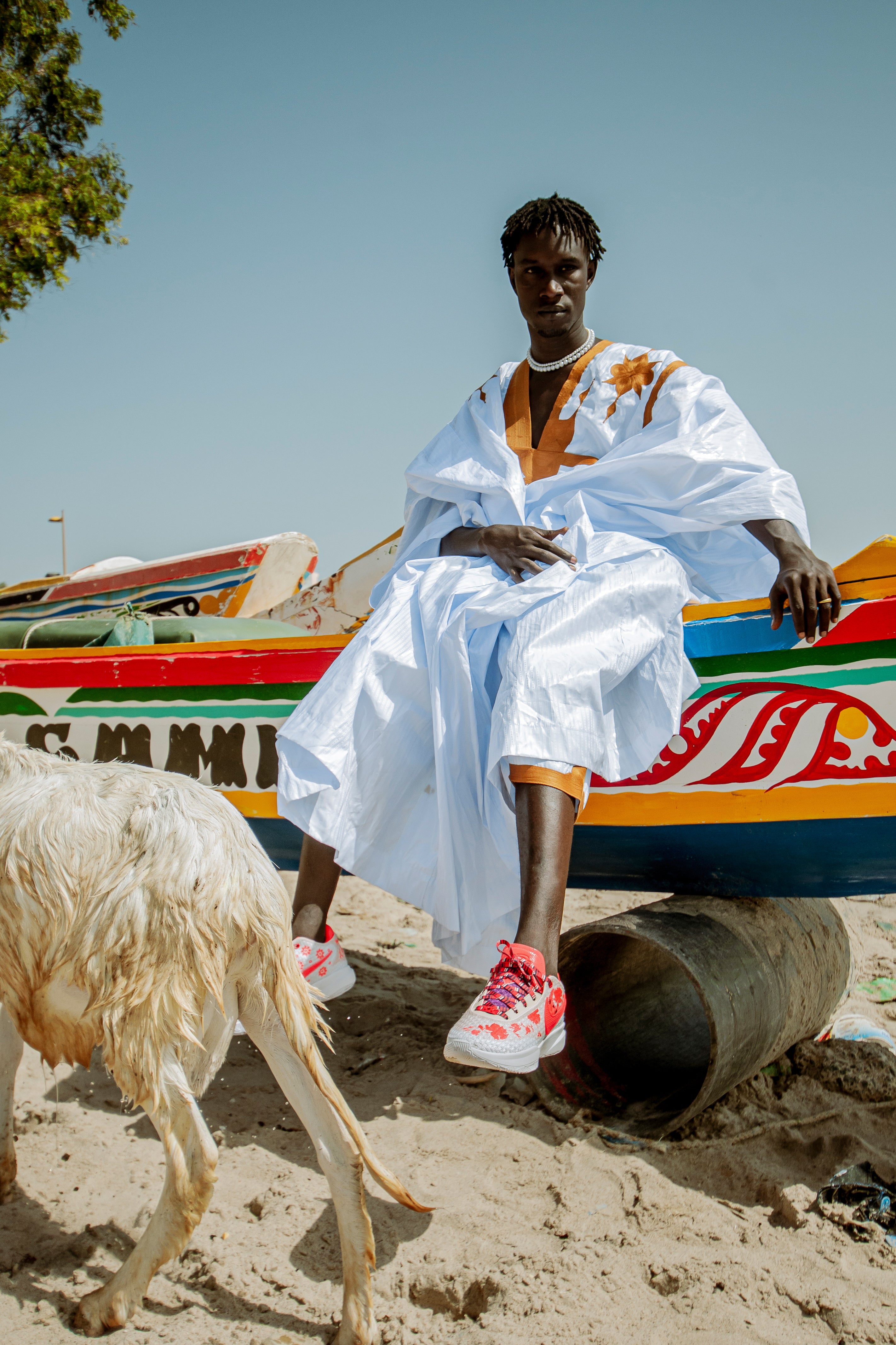 Person in a white robe sitting on a colorful boat with a goat nearby wearing basketball sneakers.