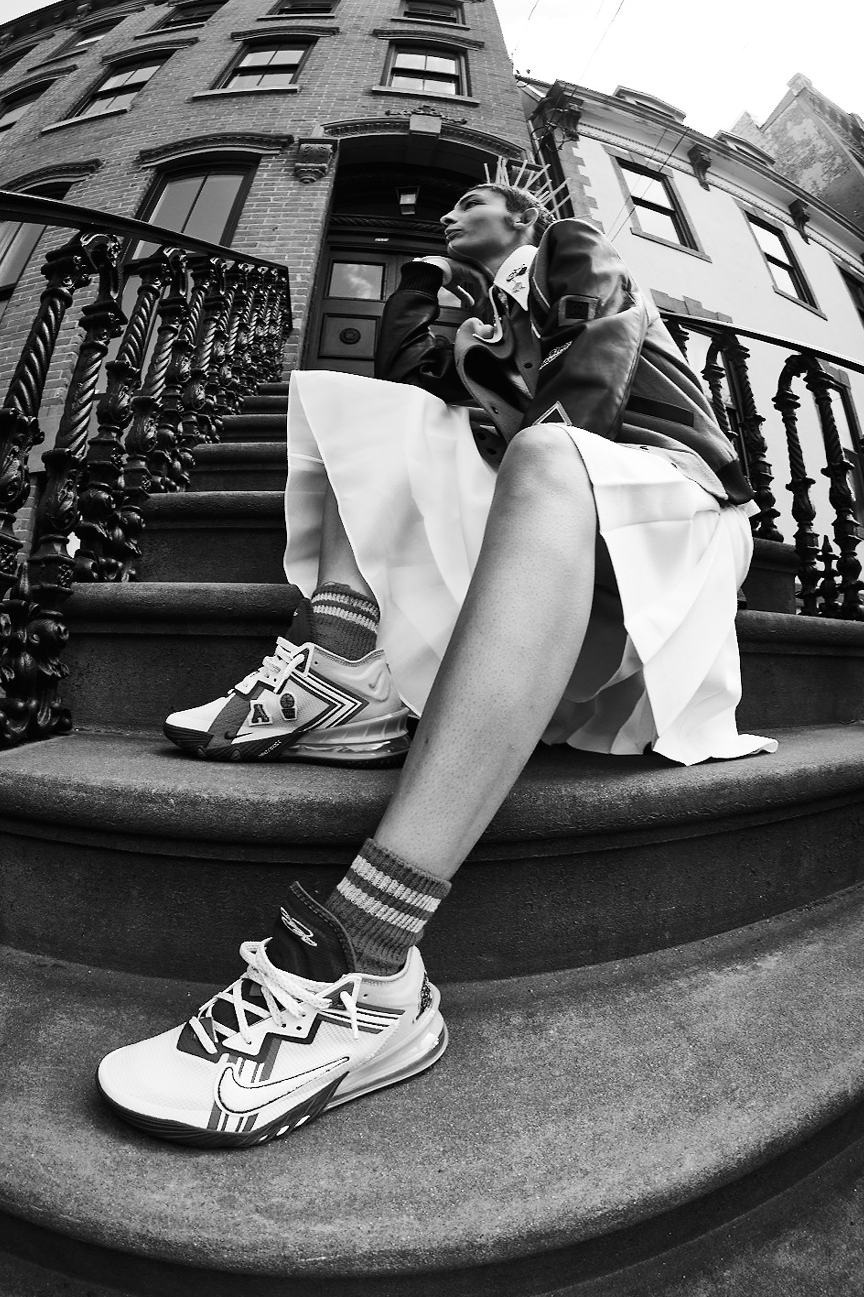 Person sitting on steps wearing a varsity jacket, skirt, and sneakers with a city building in the background