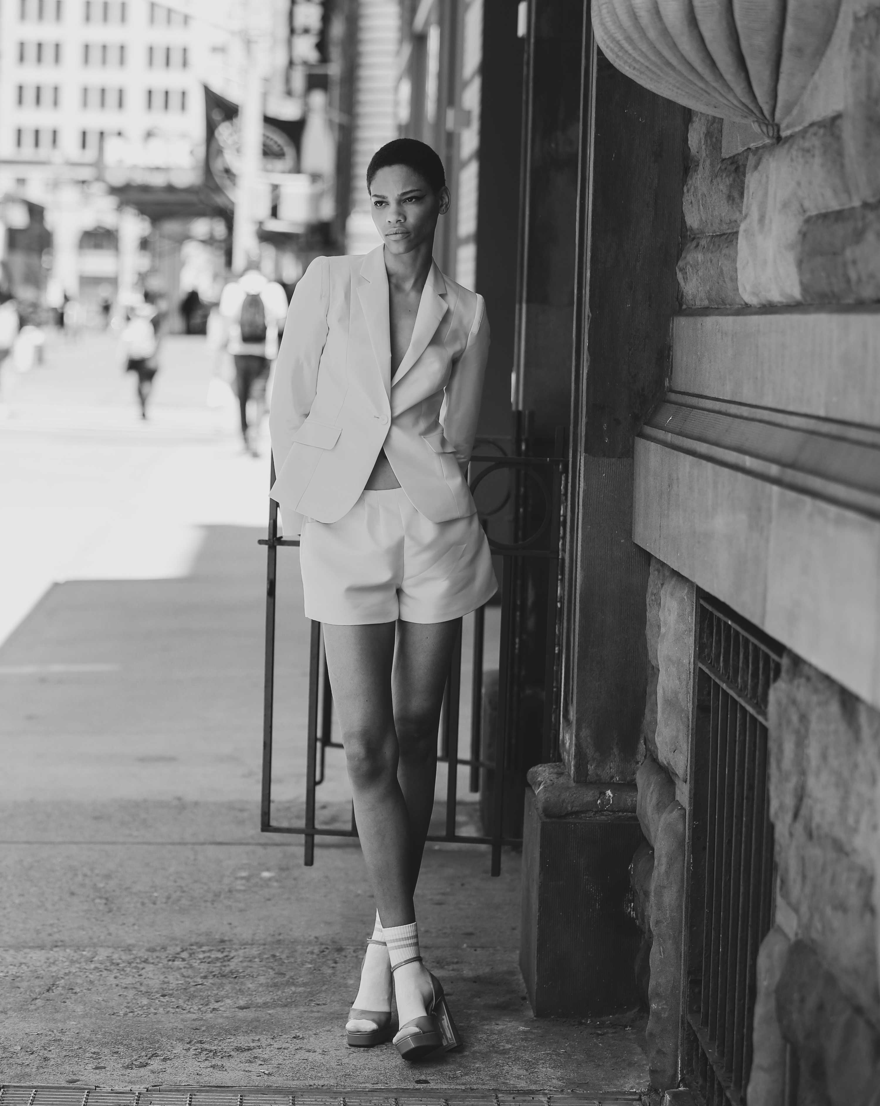 A black and white photo of a Woman wearing a coral blazer suit with shorts standing on a New York city street.