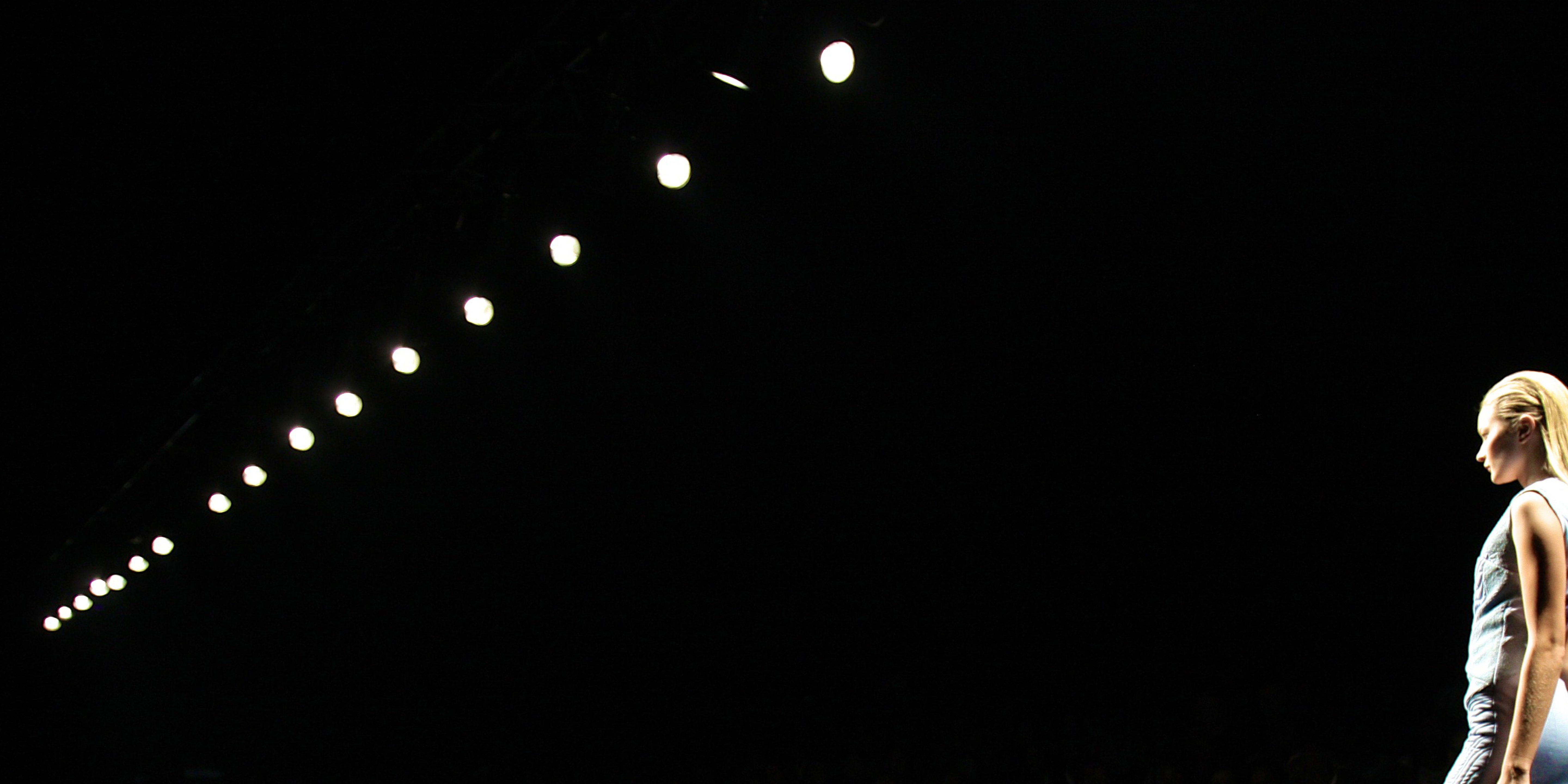 A blonde woman walks down a fashion runway in a blue quilted dress with bright lights above