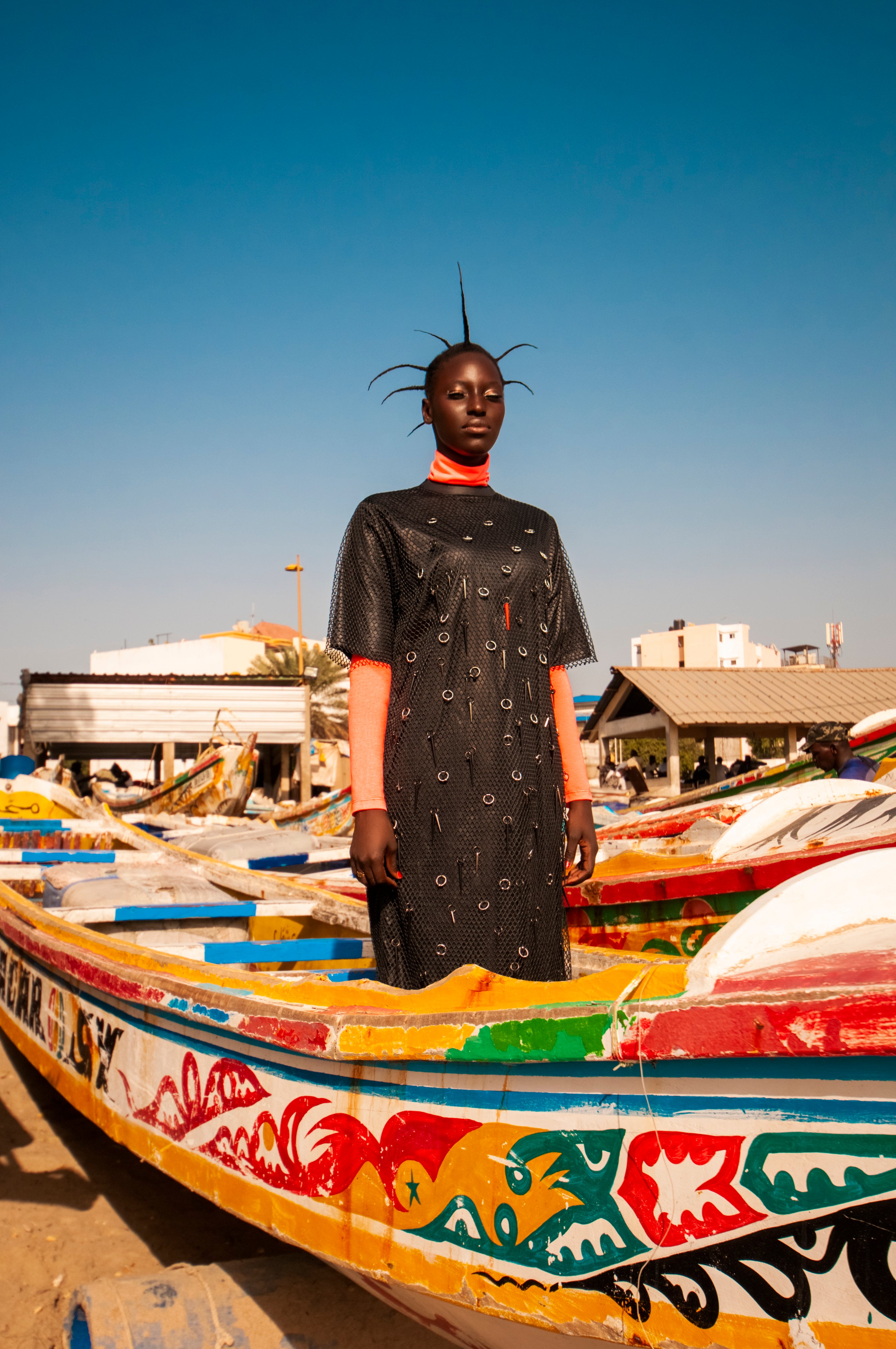 A woman model wears a black mesh dress with silver rings and neon spikes, with neon coral turtleneck by Mimi Plange on a boat on a beach in Dakar Senegal
