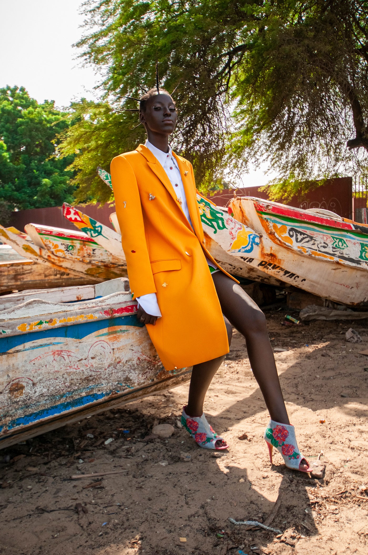 A model from Dakar Senegal wear a bright mustard yellow asymmetrical blazer and white shirt with plaid buttons against a boat on a beach