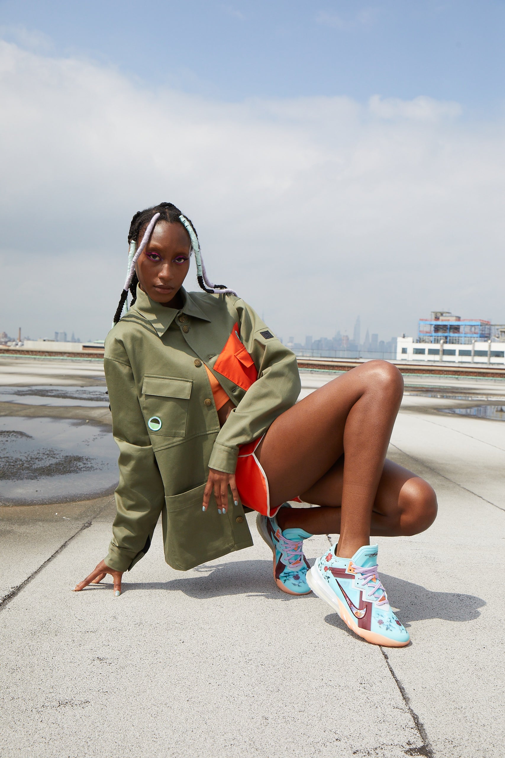 Model wearing a colorblack green coat and colorful LeBron Jamex x Mimi Plange Nike basketball sneakers on a concrete surface with a cityscape in the background