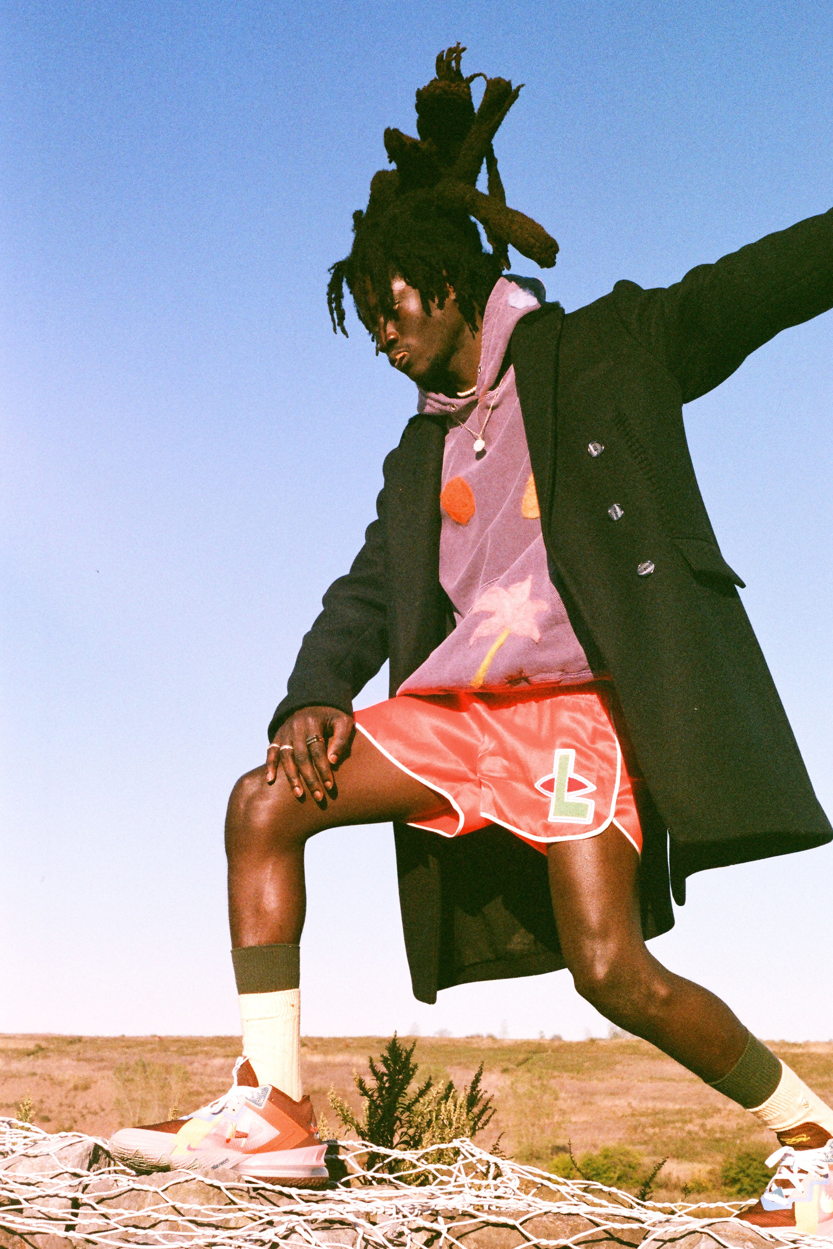 Person in a black coat and coral shorts and purple hoody with felting, standing outdoors against a blue sky.