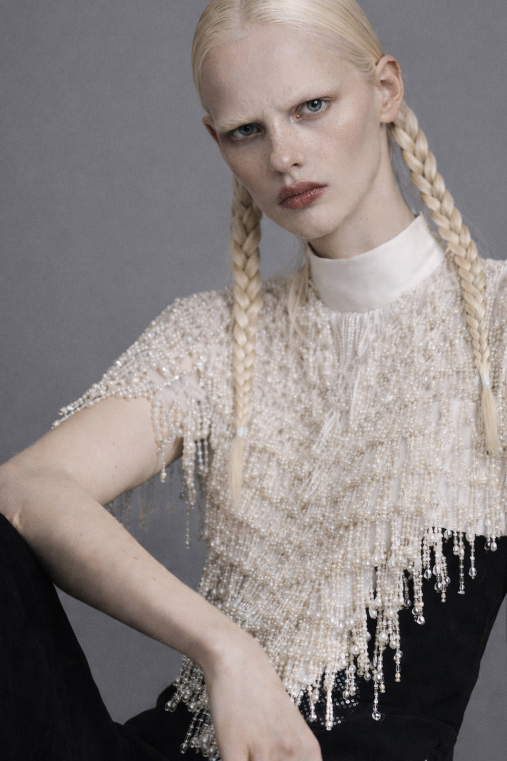 Woman with braided hair wearing a beaded top with pearls against a gray background