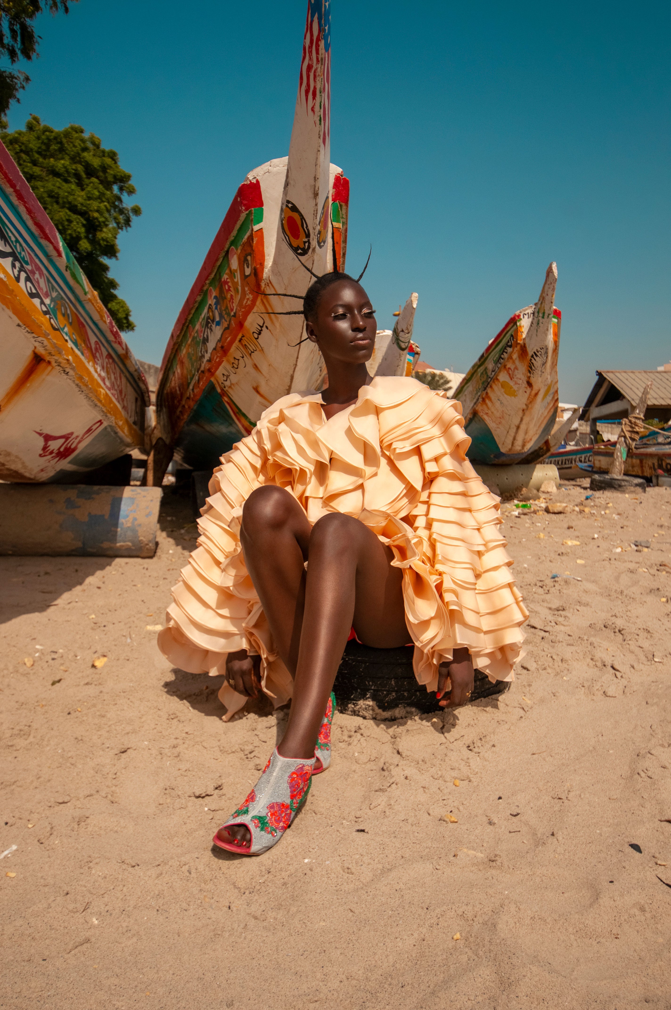 A woman wears an oversize silk ruffle coat by Mimi Plange sitting on sand on a beach in Dakar Senegal, wearing seed beaded shoes by Manolo Blahnik x Mimi Plange