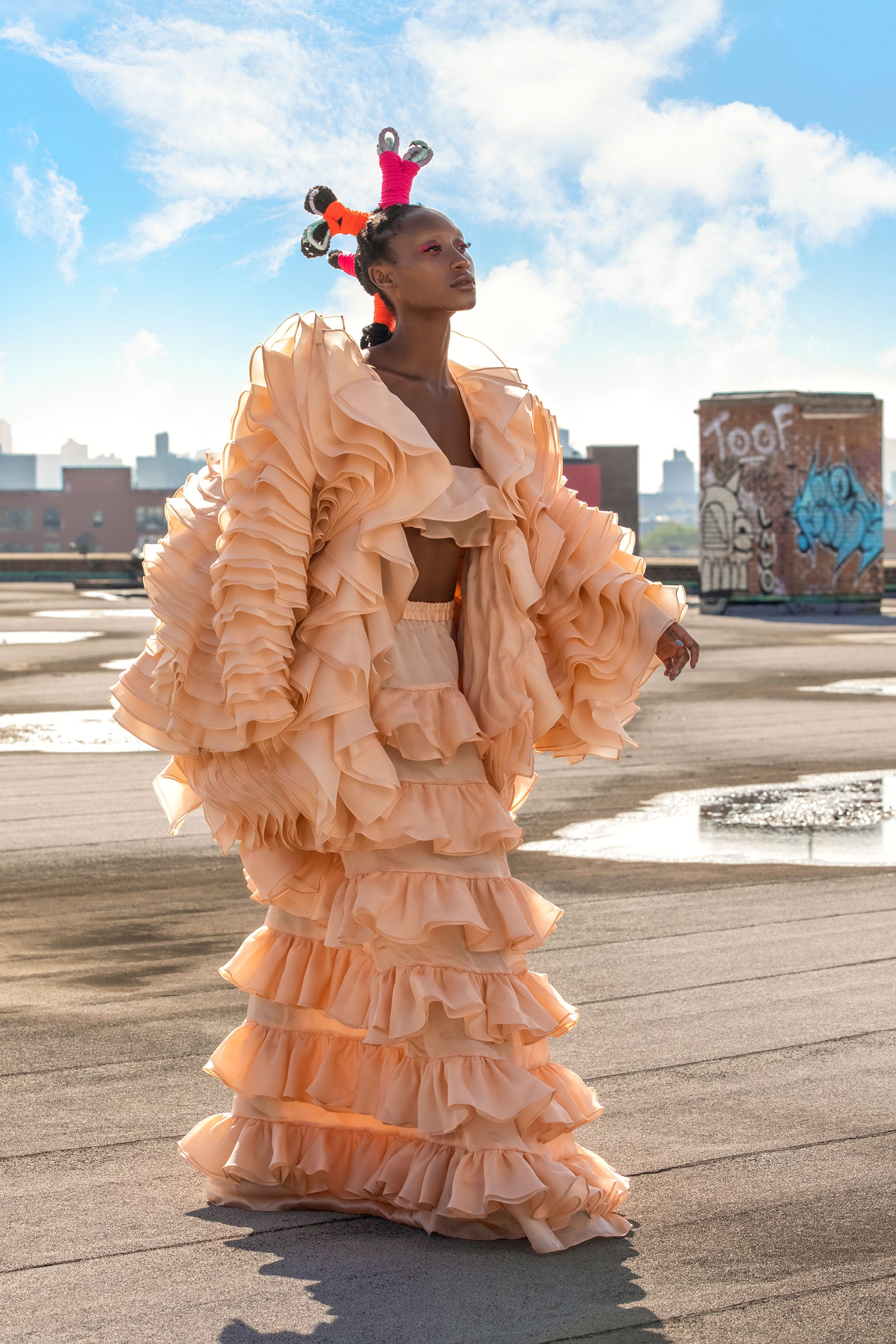 A model wears a peach colored ruffle coat and maxi ruffle skirt on a rooftop in Manhattan walking on her own runway by Mimi Plange