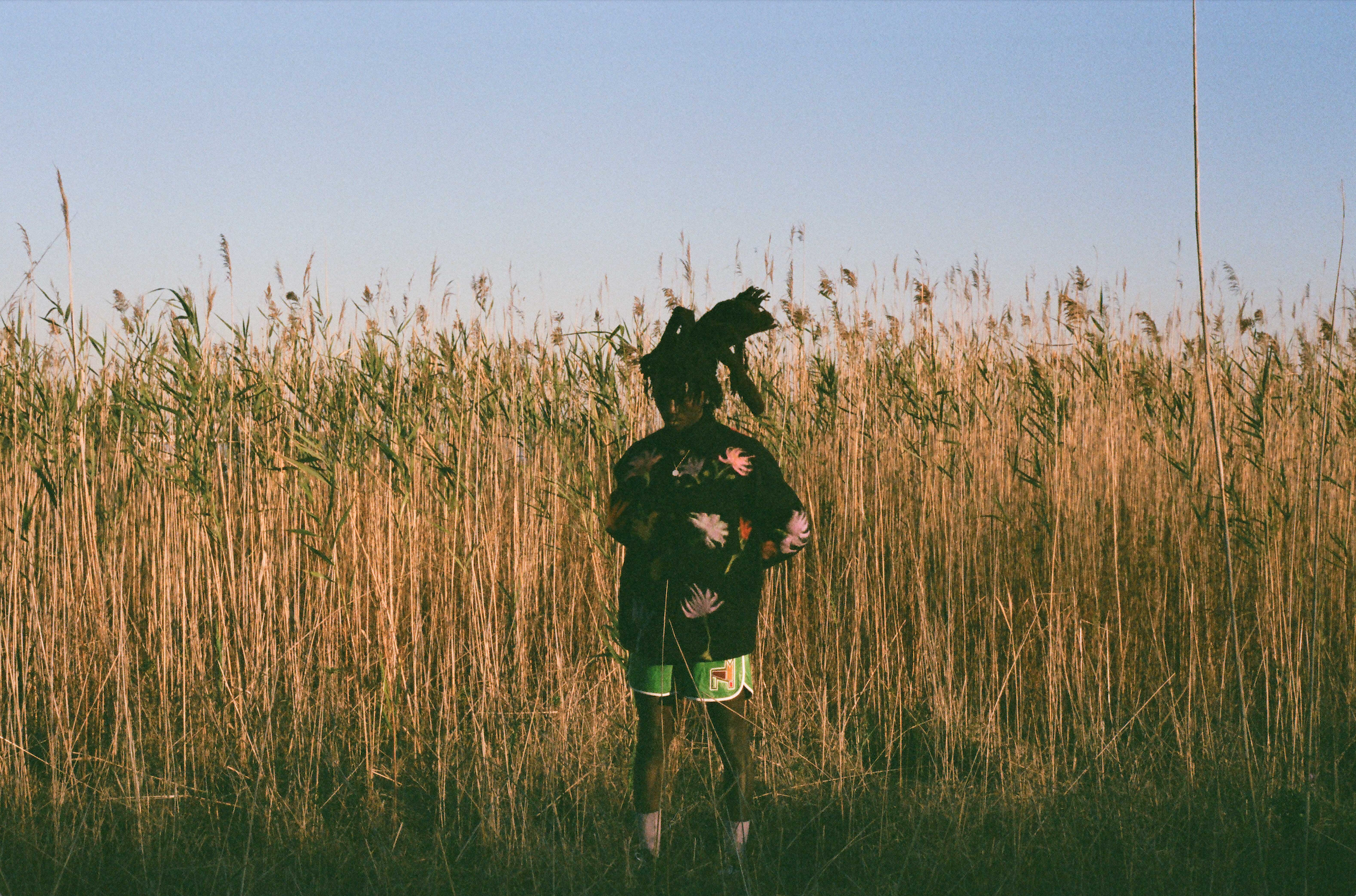 Young man wearing a hand felted jacket and leather short with patch, standing in tall grass with a clear blue sky