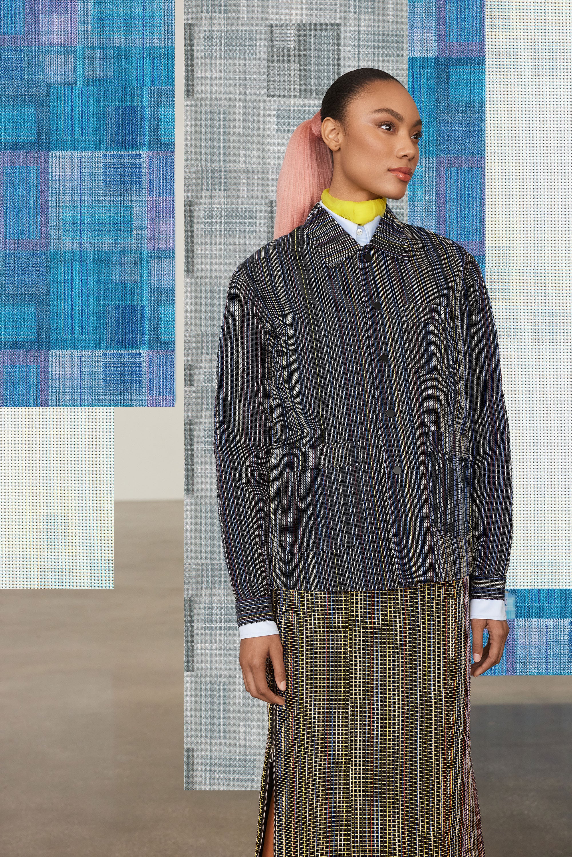 Model with pink hair wearing a textured striped jacket and plaid pencil skirt against The Shade Store x Mimi Plange window shades and treatments