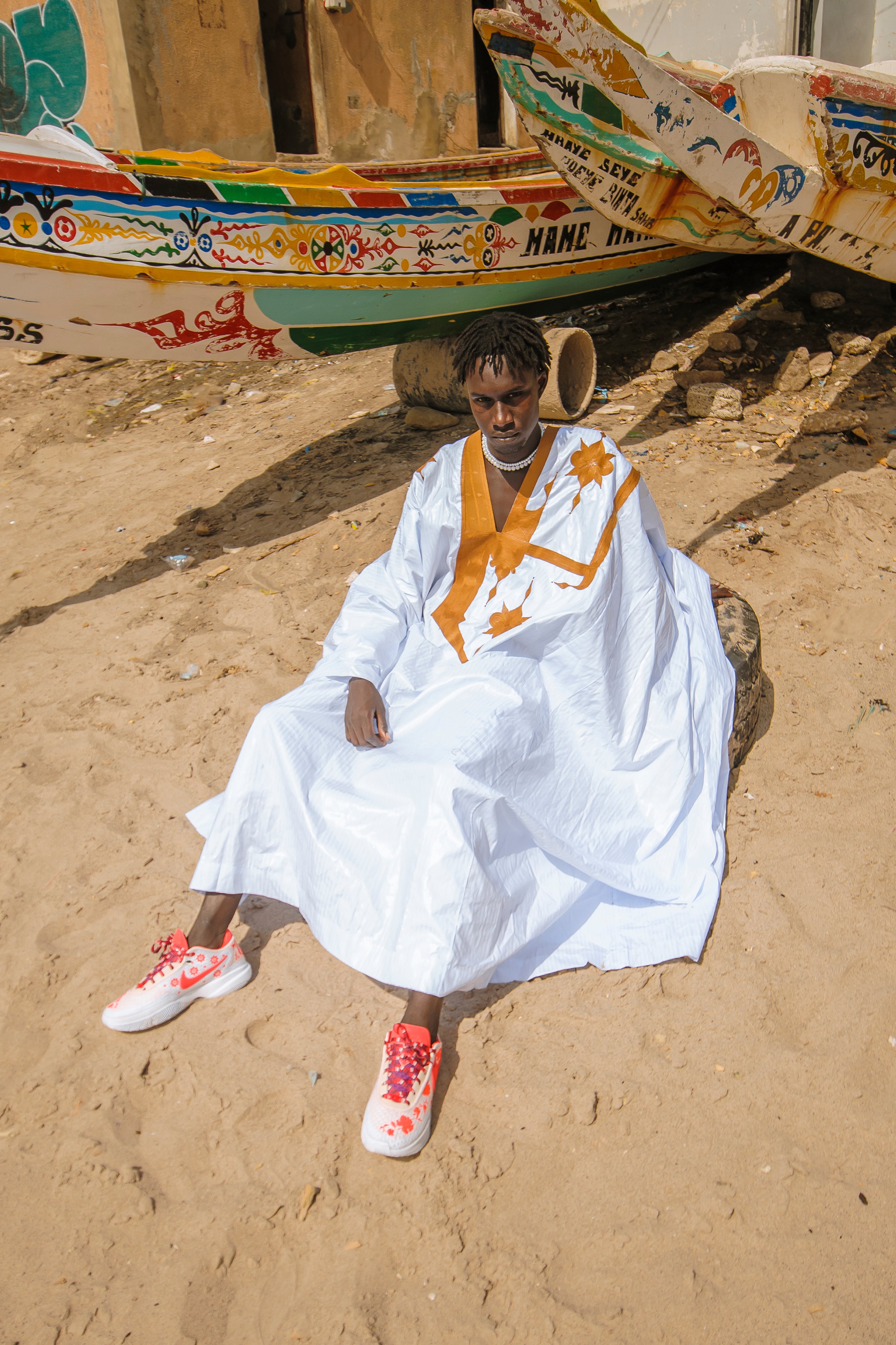 Senegelese model  in a white caftan with orange patterns sitting on sand next to a colorful boat wearing basketball sneakers.