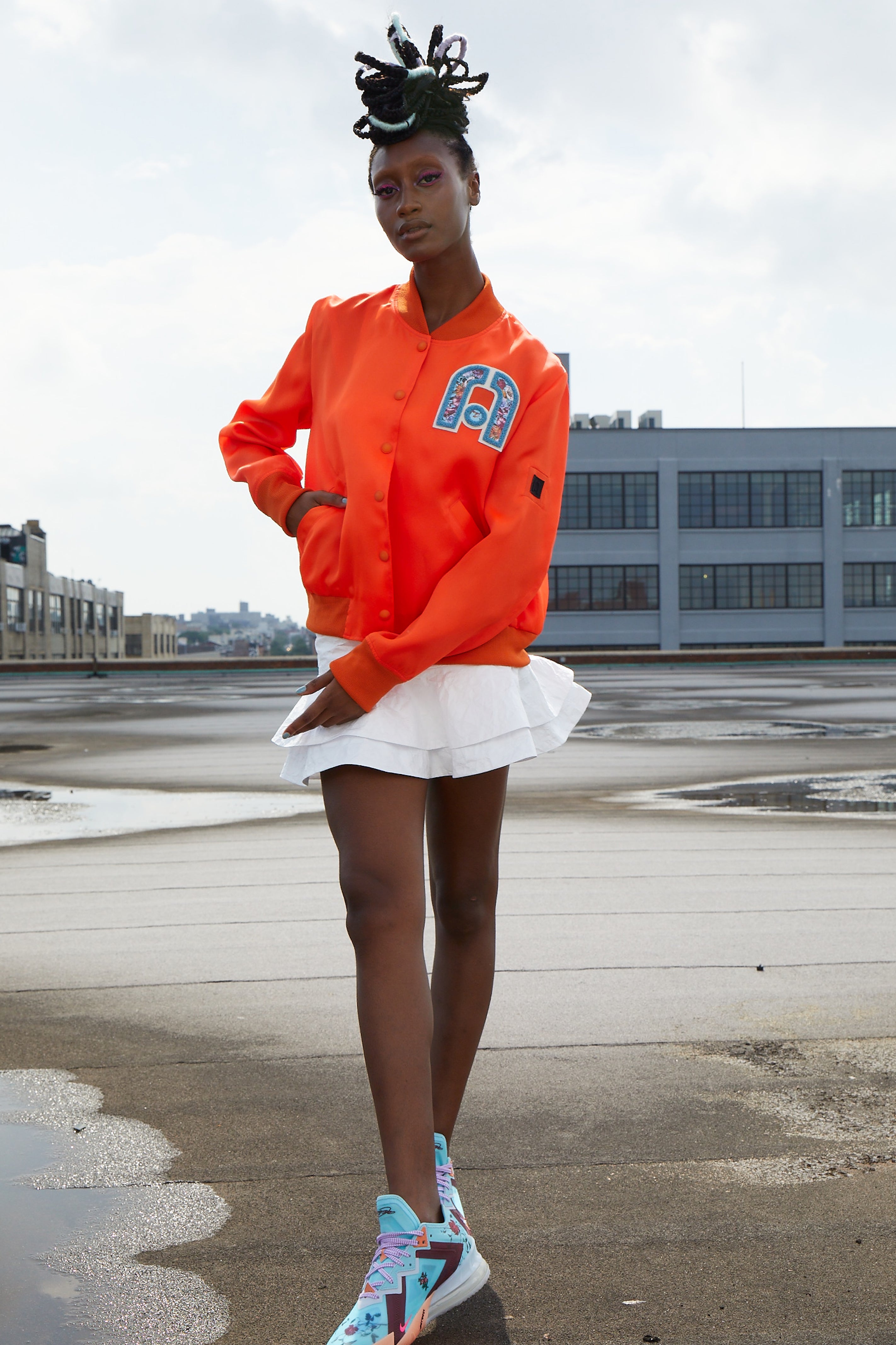 Woman wearing an orange varsity jacket with a logo, white skirt, and colorful basketball shoes on a rooftop.