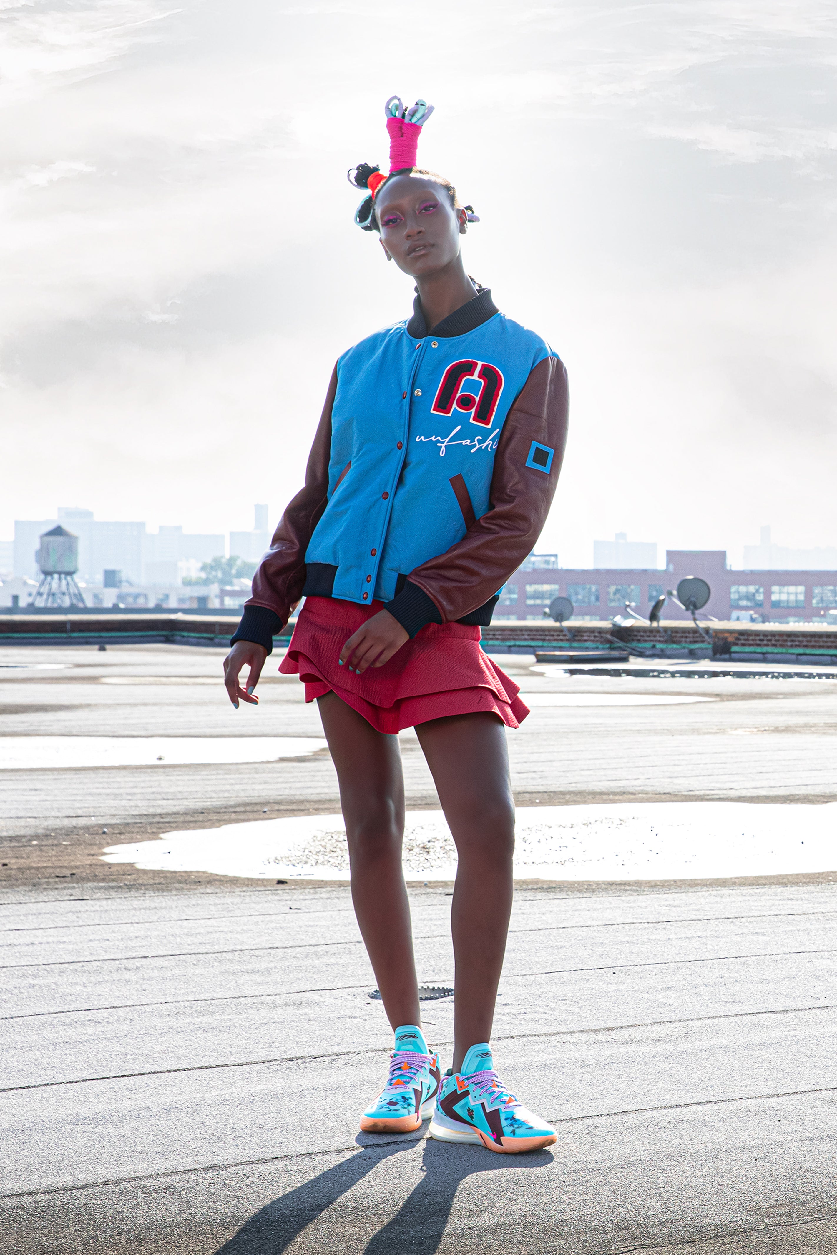 Model wearing a colorful outfit with a blue and burgandy varsity jacket and a red skirt on a rooftop.