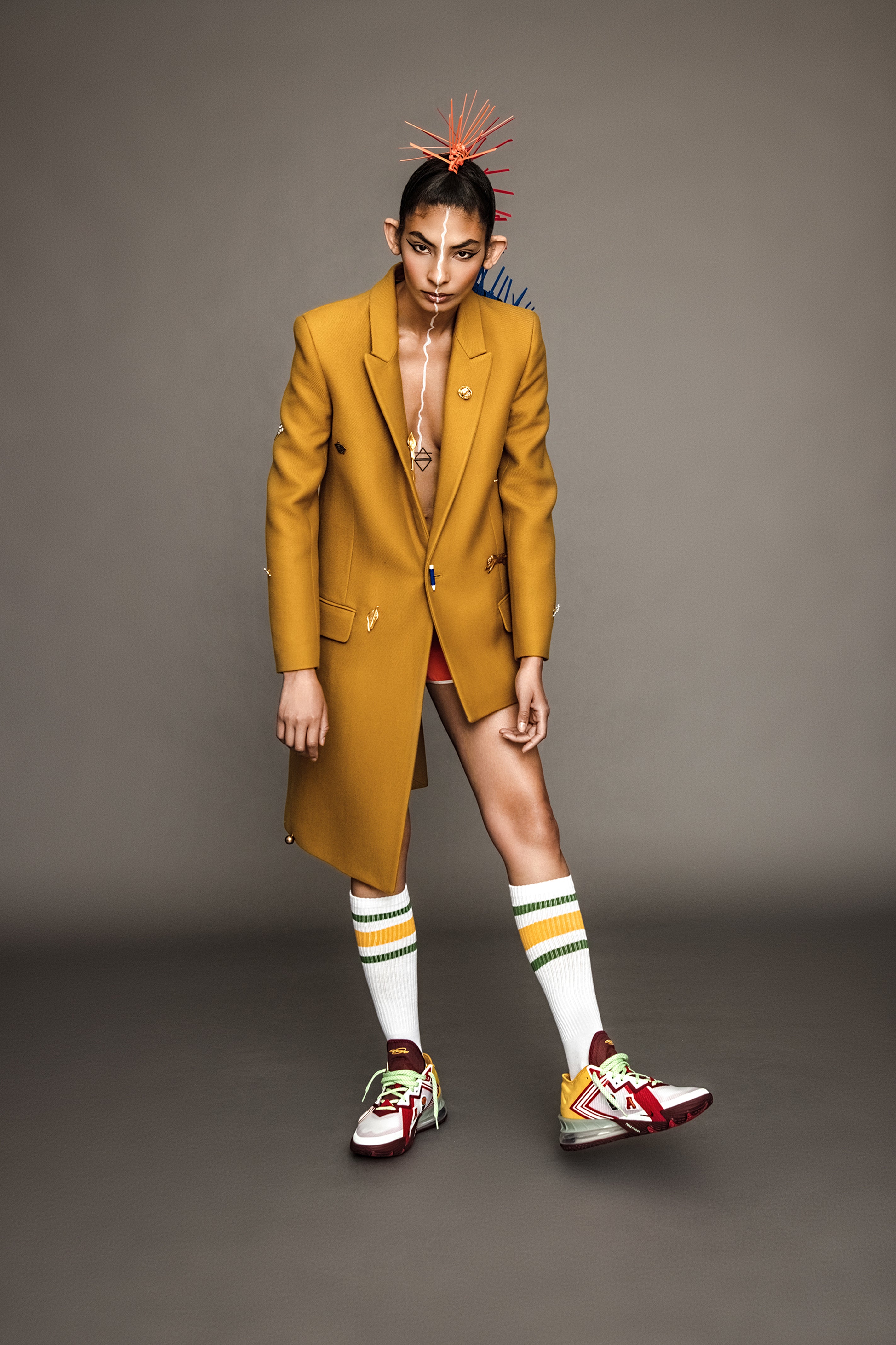 A woman wearing a mustard yellow blazer coat with colorful socks and basketball shoes on a gray background