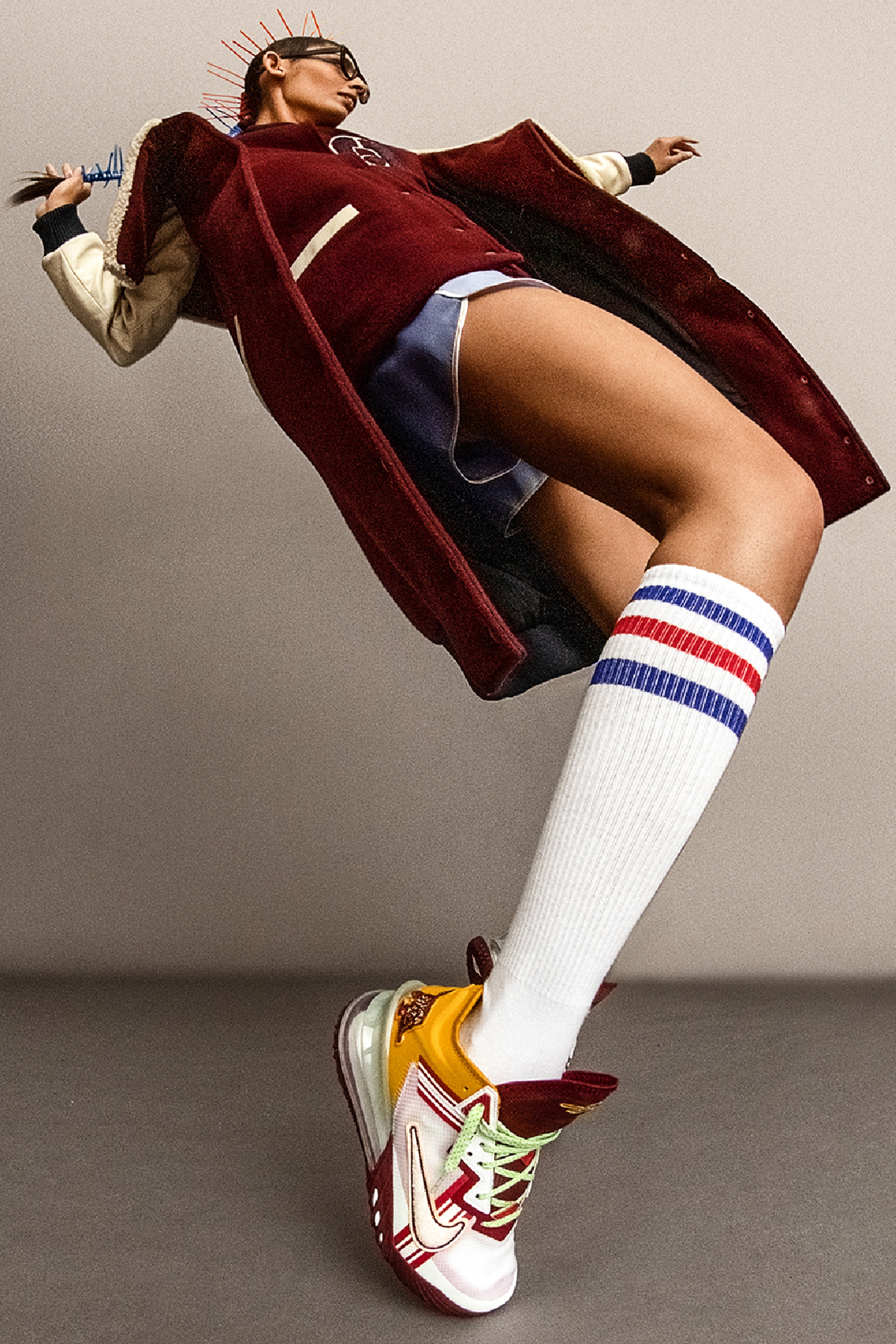 Model wearing a long burgundy varsity jacket with white knee-high socks with red and blue stripes, and colorful basketball sneakers on a plain background.