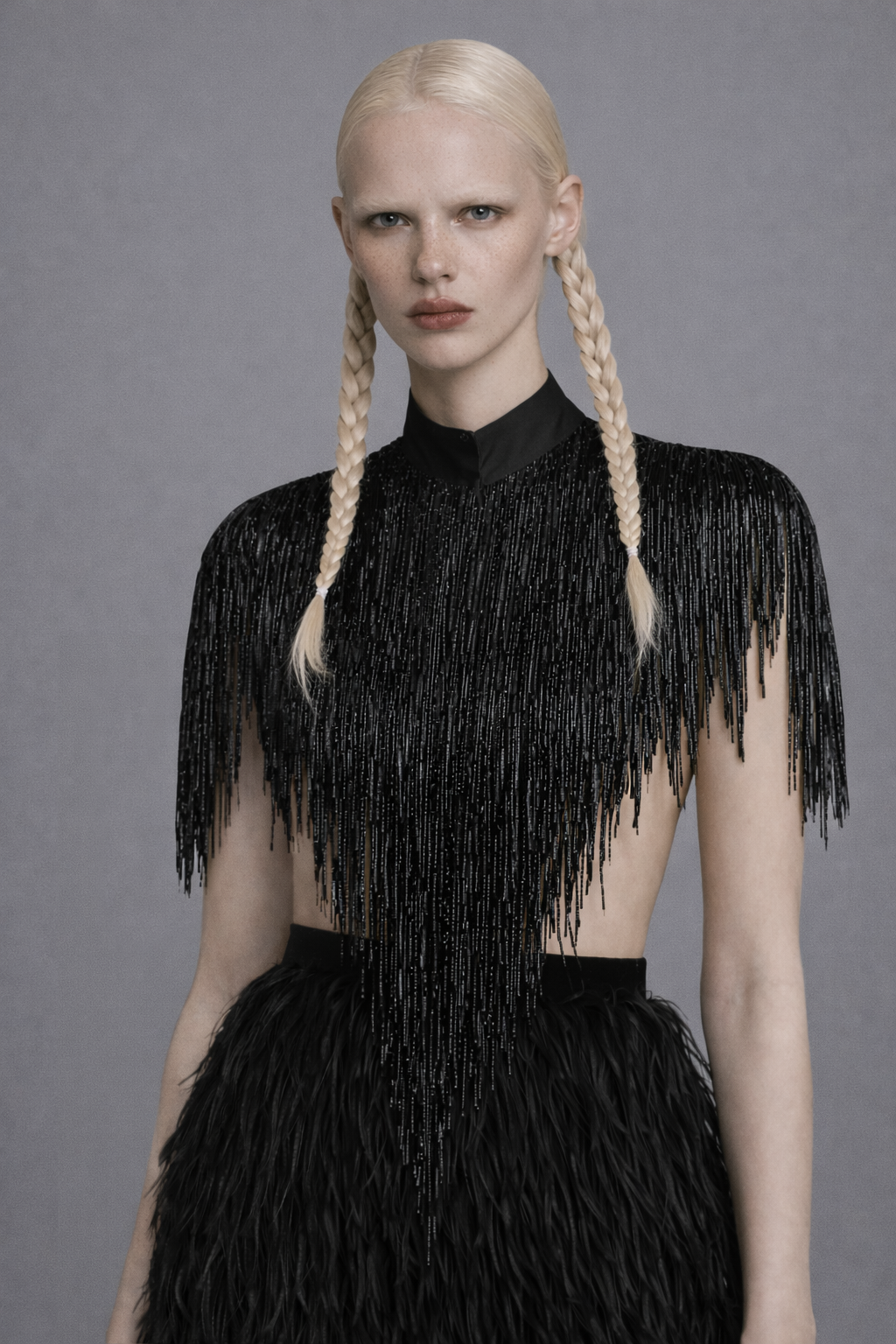 Model wearing a black fringe crop cape top and ostrich feather skirt against a gray background