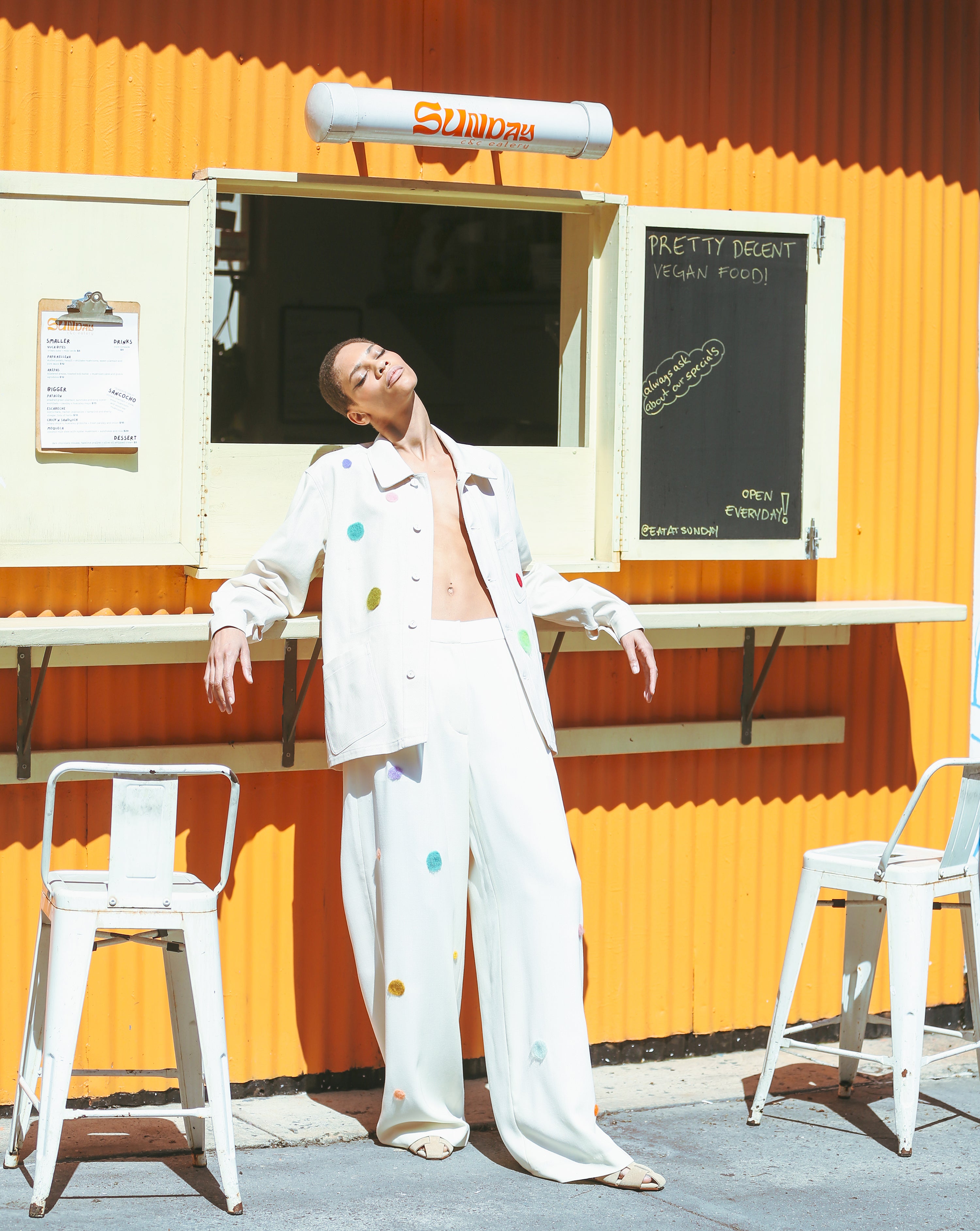 Woman in a natural canvas barn jacket with hand felted dots and trousers with needle felting and colorful polka dots standing in front of a small food truck.