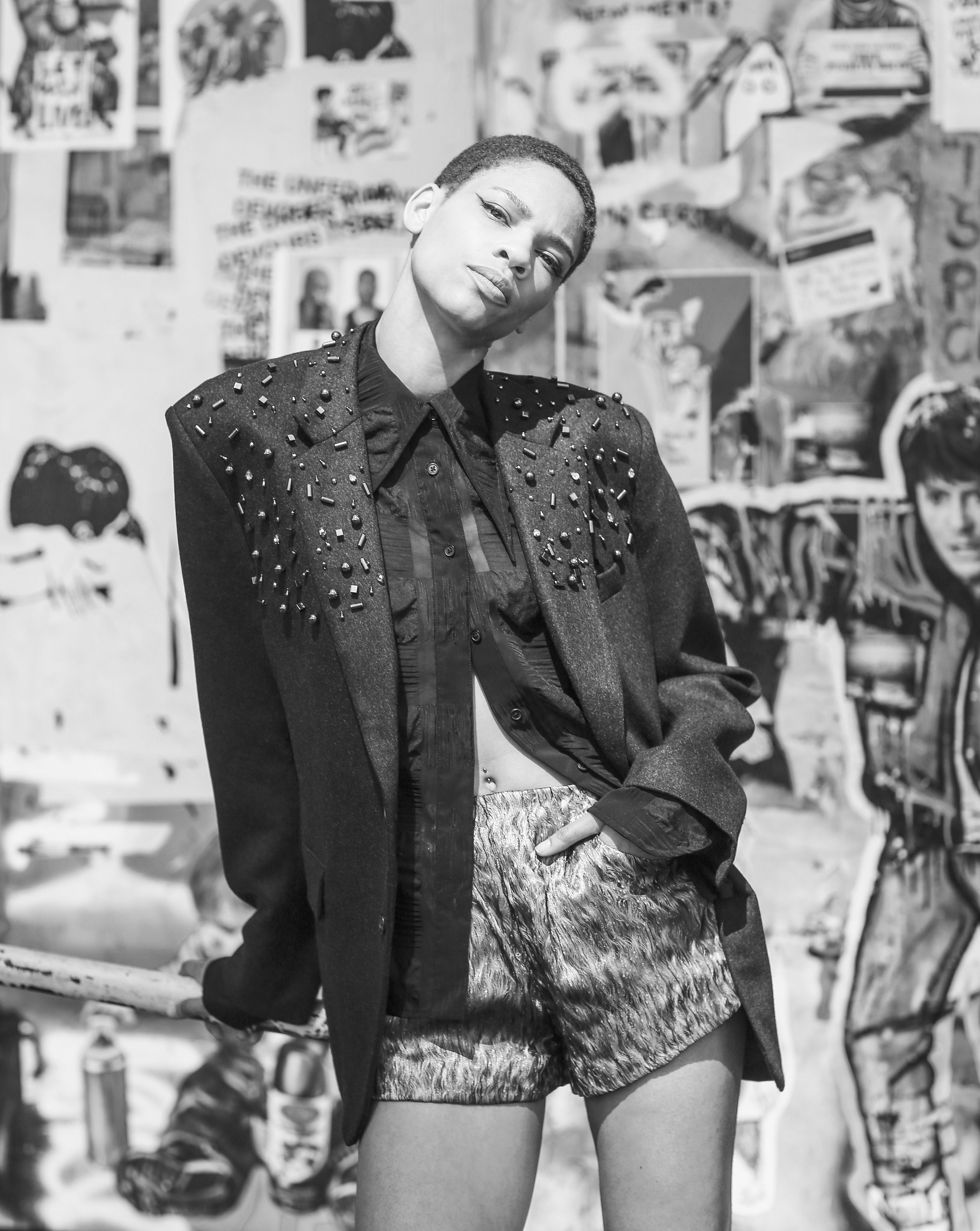 A model wears Mimi Plange metal and rhinestone embroidery blazer with silk woven shirt and shorts on a graffiti background