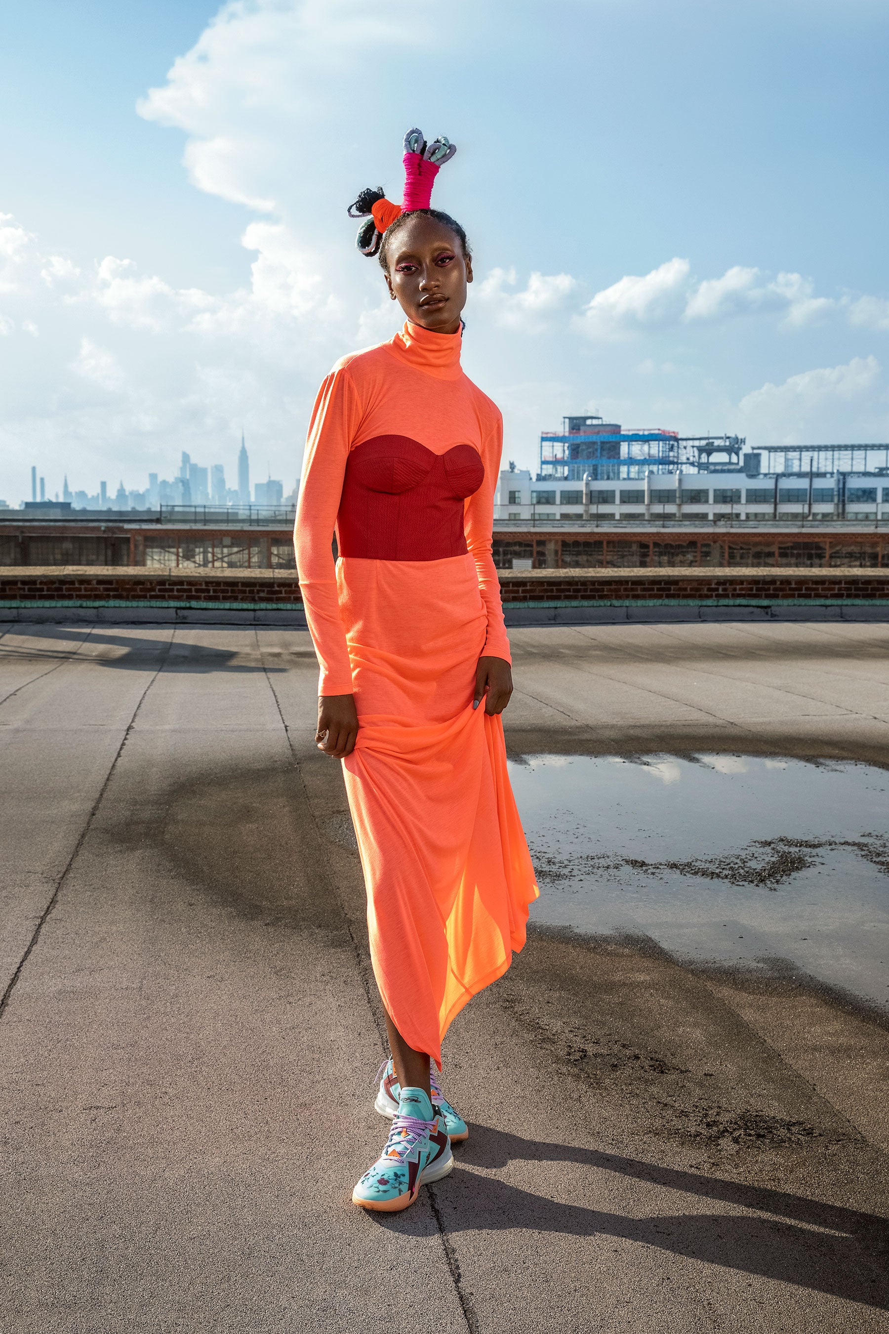 A model wears a neon coral orange turtle neck dress with a red bustier and Daughters Nike Basketball sneakers with sculpted hair.